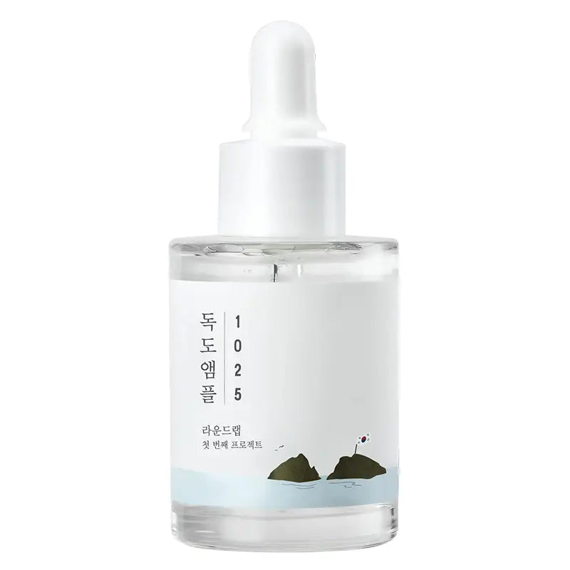 Clear glass bottle with white dropper cap containing ROUND LAB 1025 Dokdo Ampoule Serum 45g, featuring minimalist label with Korean text and a small illustration of two islands with a flag.