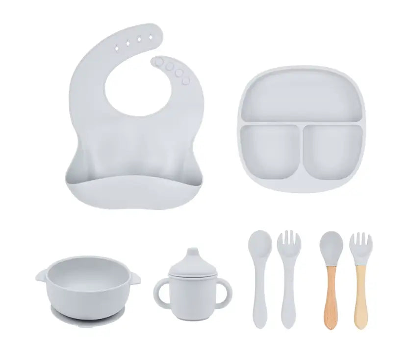 Collection of baby feeding utensils including a bib, plate, bowl, cup, and spoons on a white background.