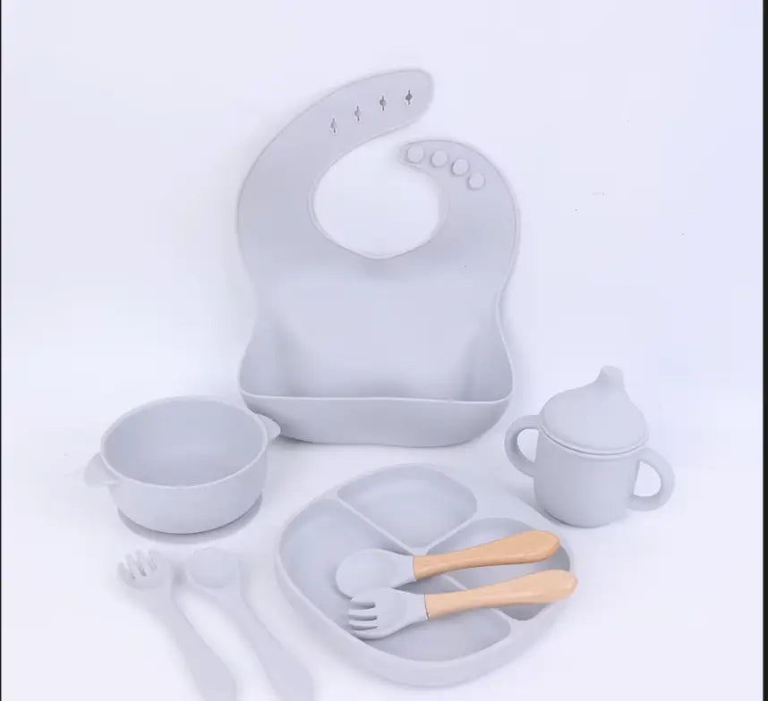 Set of baby feeding utensils including a bib, bowl, spoon, and cup on a light gray background.