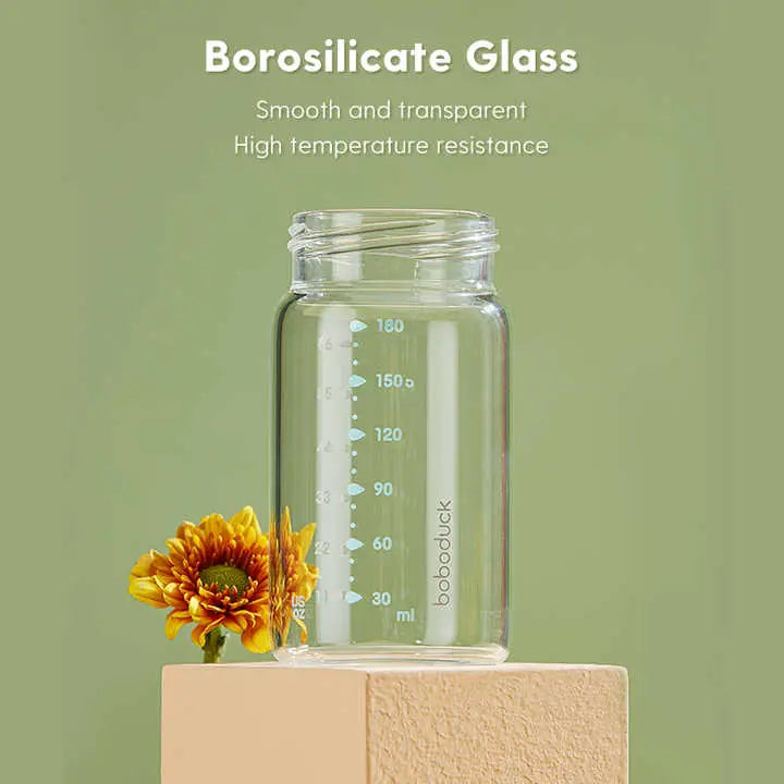Clear borosilicate glass baby bottle with measurement markings on a beige pedestal and yellow flower, highlighting smooth transparency and high temperature resistance for All-in-One Wide-Neck Glass Baby Milk Storage Bottle | Pump Directly into Bottle Store in Refrigrator and Feed Directly from Glass Bottle| BPA-Free | Glass Breas.