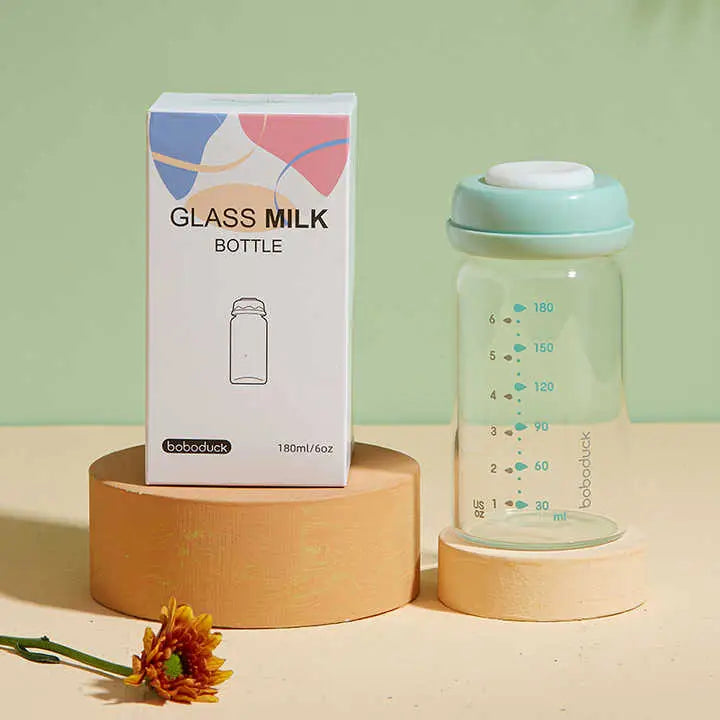 All-in-One Wide-Neck Glass Baby Milk Storage Bottle | Pump Directly into Bottle Store in Refrigrator and Feed Directly from Glass Bottle| BPA-Free | Glass Breas displayed next to its packaging box on a wooden stand with a flower nearby on a neutral background
