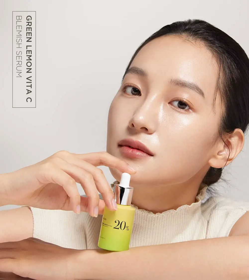 Woman with clear skin holding a green and yellow bottle of Anua Green Lemon Vitamin C Blemish Serum 20ml against a light background with product name text visible.