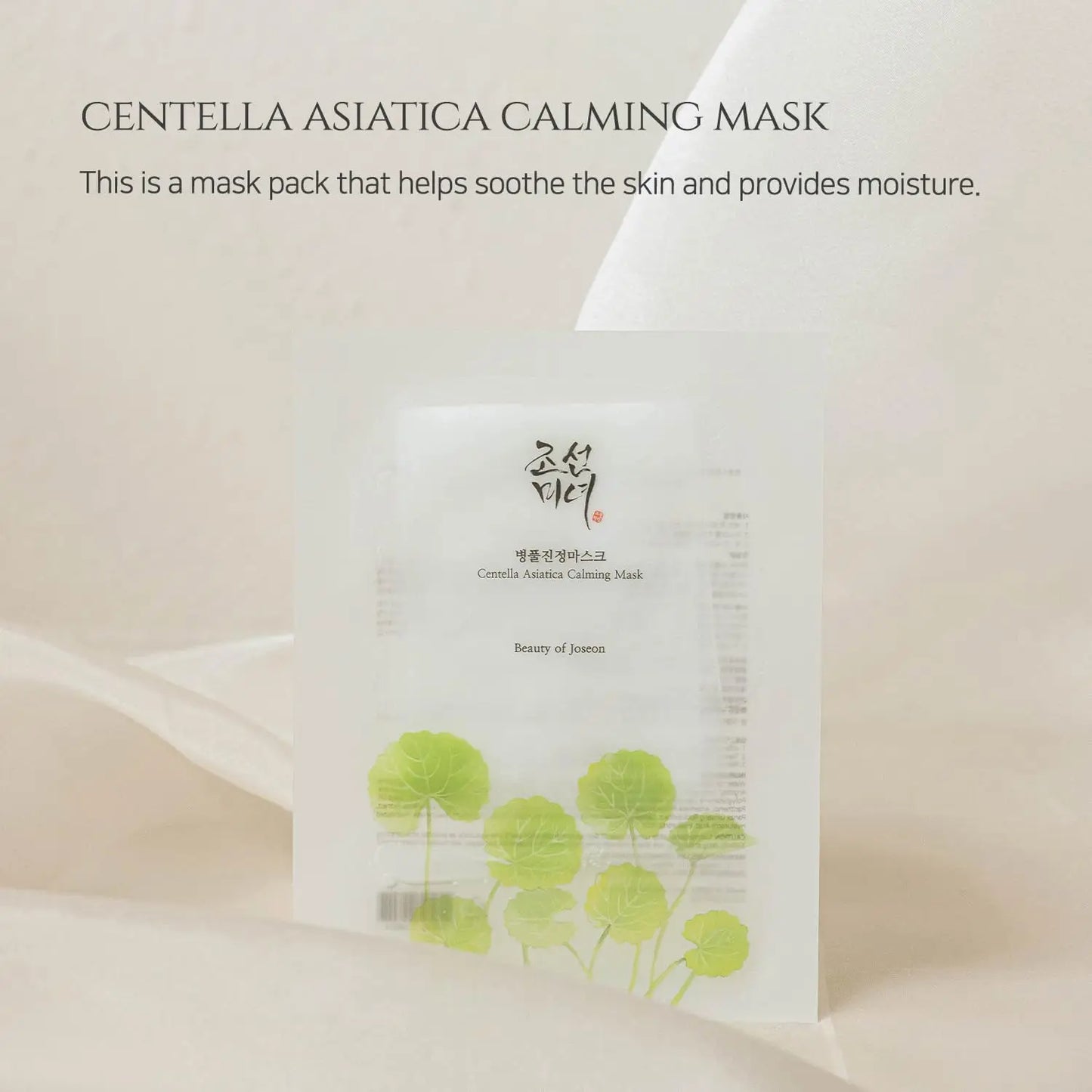 Minimalist product image of Beauty of Joseon Centella Asiatica Calming Mask pack standing with green centella asiatica leaves illustration on a soft beige fabric background.