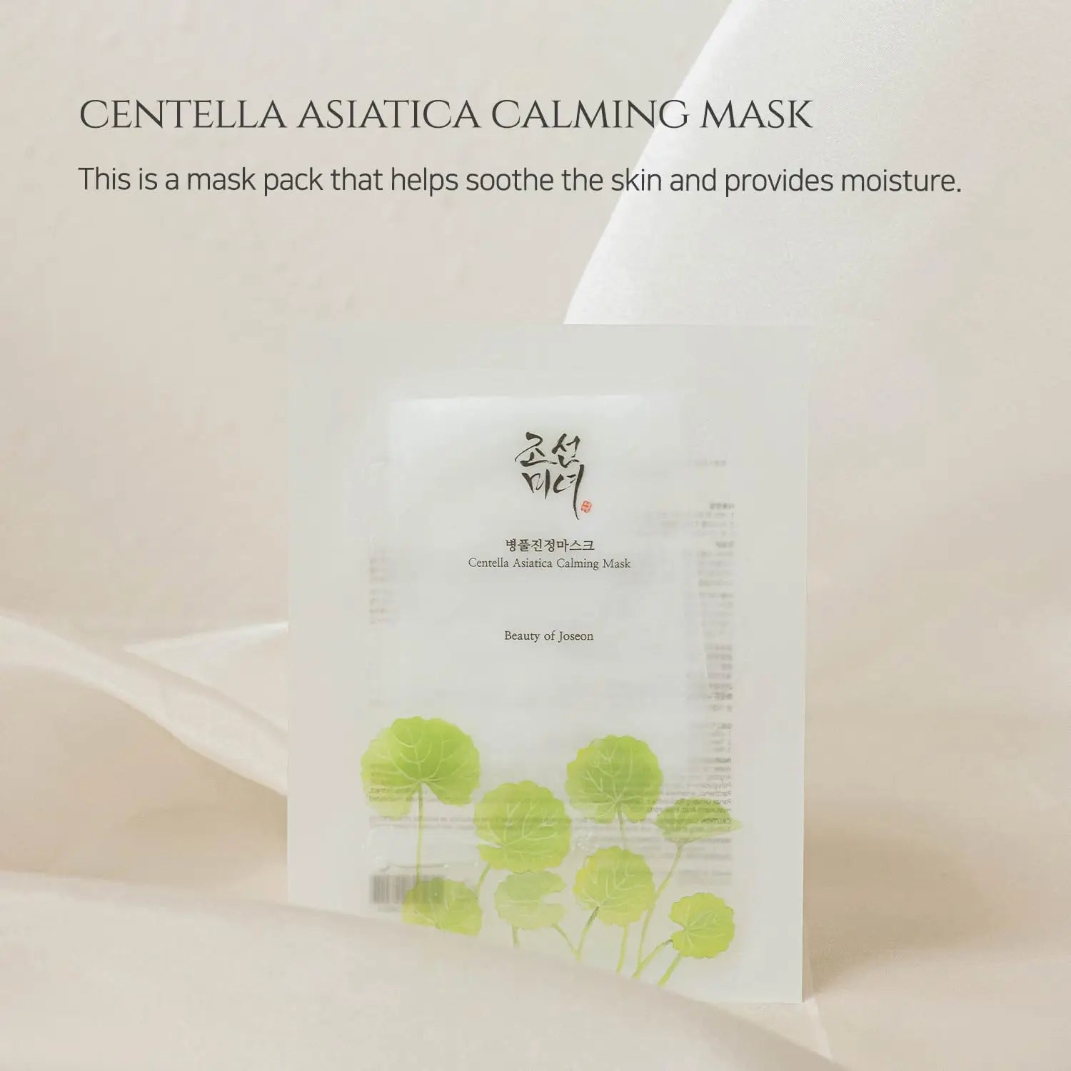 Minimalist product image of Beauty of Joseon Centella Asiatica Calming Mask pack standing with green centella asiatica leaves illustration on a soft beige fabric background.