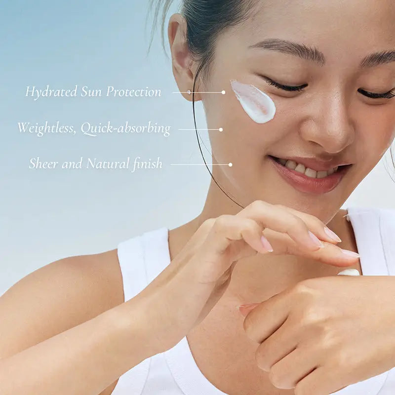 Close-up of a smiling woman applying Beauty of Joseon Relief Sun Aqua-Fresh : Rice + B5 sunscreen with visible smooth white cream on her cheek, highlighting hydrated sun protection, quick absorption, and sheer natural finish.