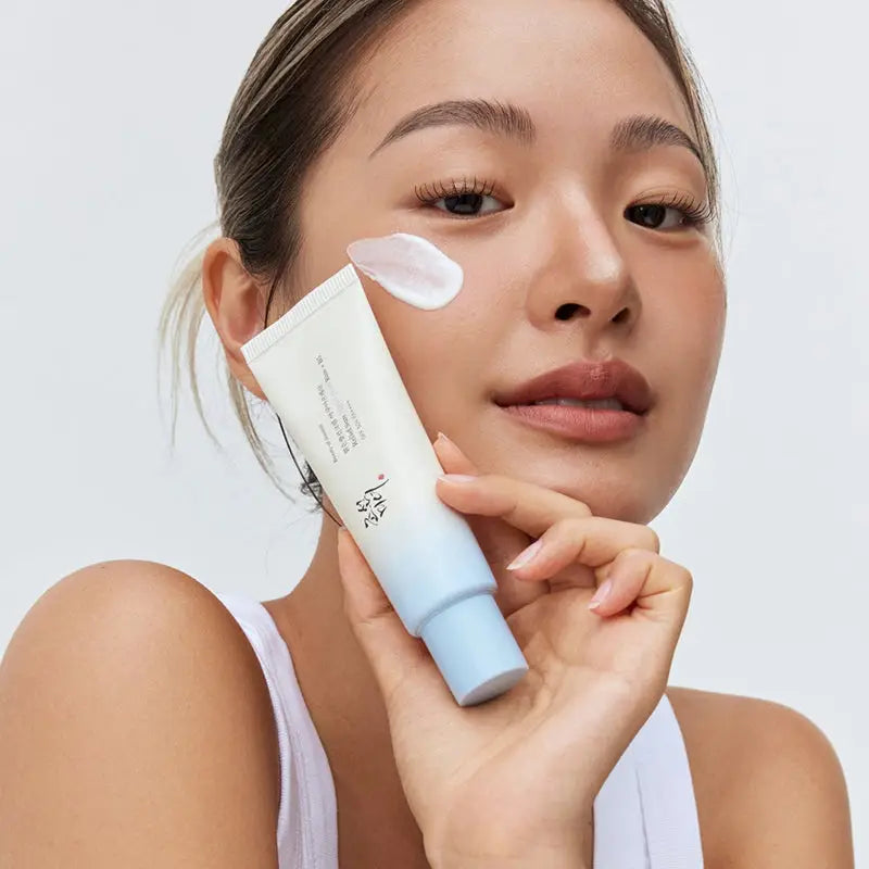 Woman applying Beauty of Joseon Relief Sun Aqua-Fresh : Rice + B5 sunscreen cream on her cheek, holding a white and blue tube, demonstrating skincare use.