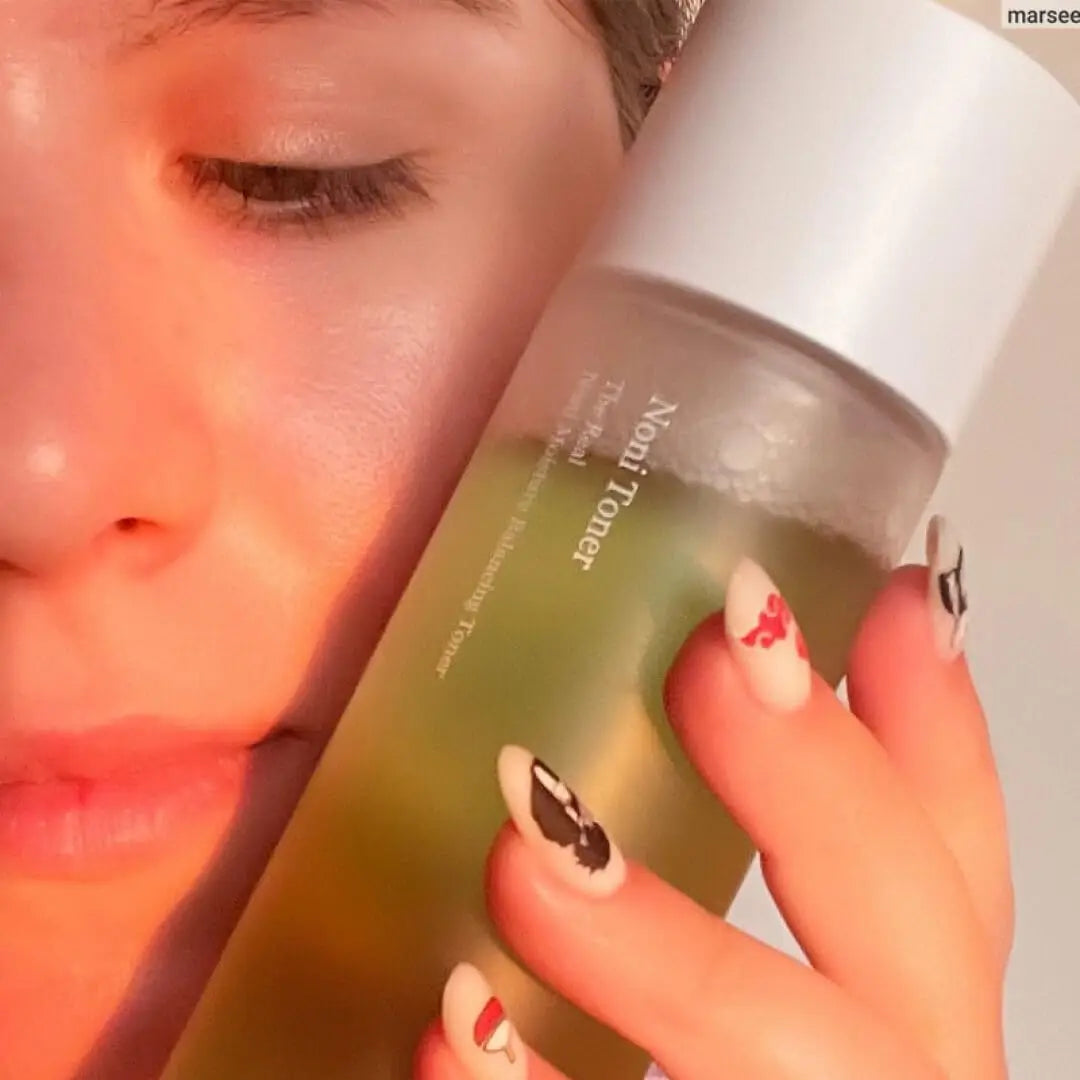 Close-up of a person gently holding a celimax Noni Moisture Balancing Toner 150ml bottle near their face, showcasing clear skin and elegant nail art.