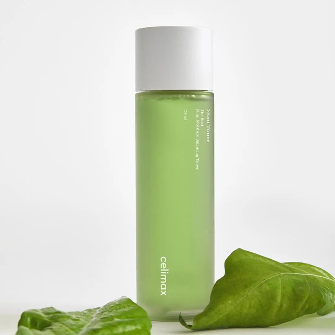 celimax Noni Moisture Balancing Toner 150ml in a clear green bottle with white cap, placed beside fresh green leaves on a white background