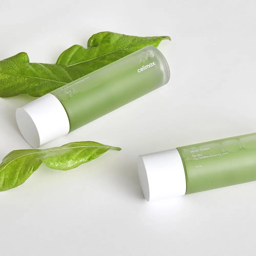 Two green bottles of celimax Noni Moisture Balancing Toner 150ml with white caps on a white surface, accompanied by fresh green leaves, highlighting a natural skincare theme.