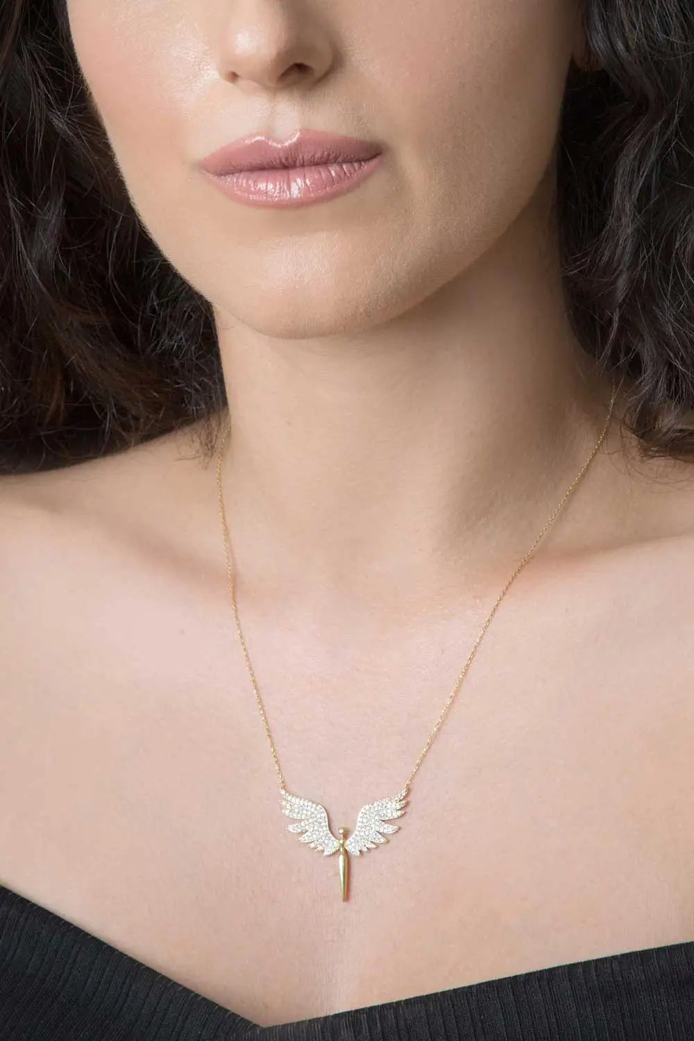Close-up of a woman wearing the Einzelstück - 14K Gold Plated Angel Necklace - Angel Gold Necklace - Gold Necklaces Gift for Women featuring a delicate gold chain and an angel wing pendant embellished with small white stones.