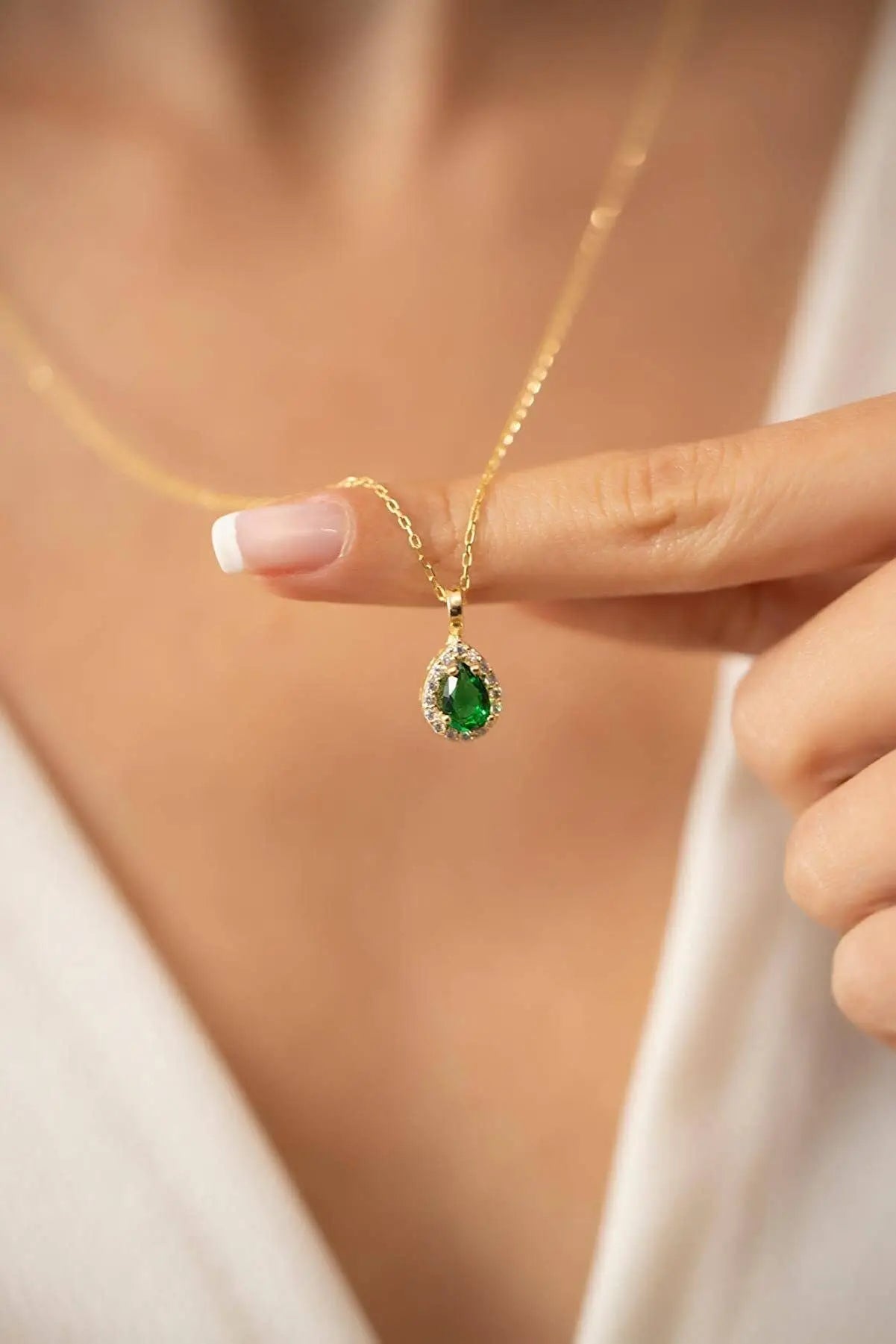 Close-up of a hand with manicured nails holding a gold chain necklace featuring a green teardrop pendant surrounded by small clear stones, showcasing Einzelstück - 14K Gold Plated Emerald Drop Necklace - Emerald water drop Necklace - Gold Necklaces Gift for Women.