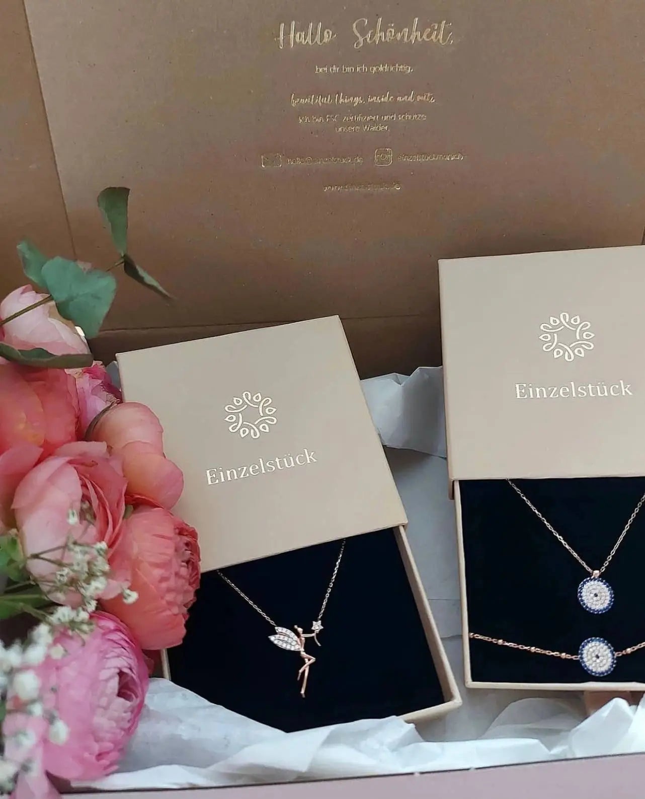 Open beige Einzelstück jewelry gift boxes containing delicate gold plated necklaces displayed with pink flowers and white decorative filler, featuring "Einzelstück - 14K Gold Plated Emerald Drop Necklace - Emerald water drop Necklace - Gold Necklaces Gift for Women" packaging.
