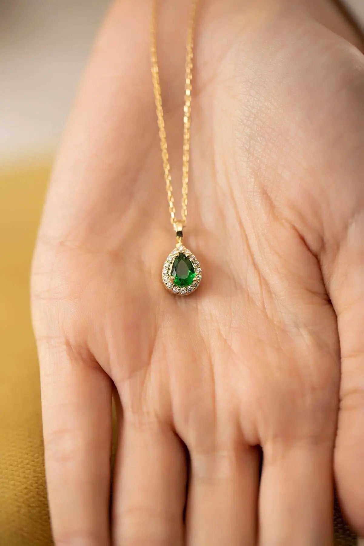 Close-up of a hand holding an Einzelstück - 14K Gold Plated Emerald Drop Necklace - Emerald water drop Necklace - Gold Necklaces Gift for Women with a teardrop-shaped green emerald pendant surrounded by small clear stones on a gold chain.