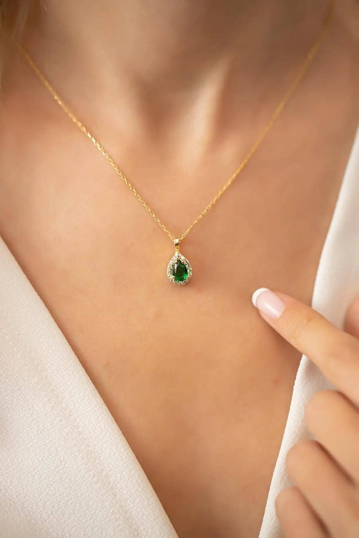 Close-up of a person wearing the Einzelstück - 14K Gold Plated Emerald Drop Necklace - Emerald water drop Necklace - Gold Necklaces Gift for Women with a teardrop-shaped green emerald pendant surrounded by small clear stones on a delicate gold chain.