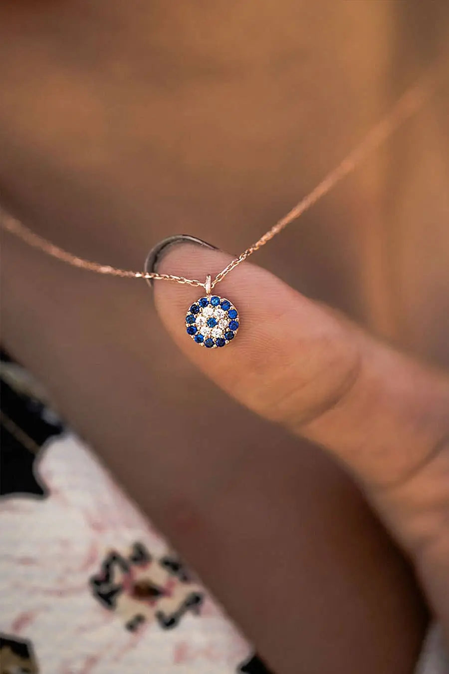 Close-up of a delicate Einzelstück - 14K Gold Plated Evil Eye Rose Gold Necklace - Evil Eye Rose Gold Necklace - Rose Gold Necklaces Gift for Women held on a finger, featuring a round pendant with blue and white stones.