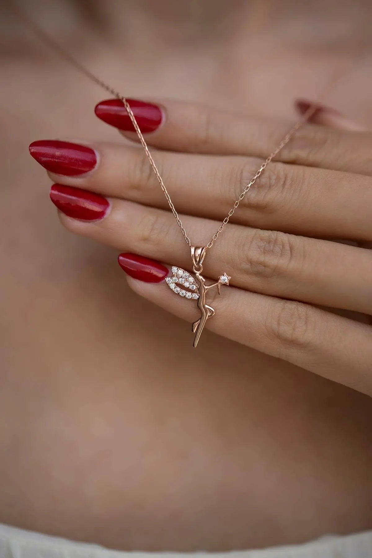 Close-up of a hand with red nail polish holding a delicate Einzelstück - 14K Gold Plated Fairy Necklace with Black Zircona stones - Fairy Gold Necklace - Gold Necklaces Gift for Women featuring a fairy pendant with sparkling wings on a fine gold chain.