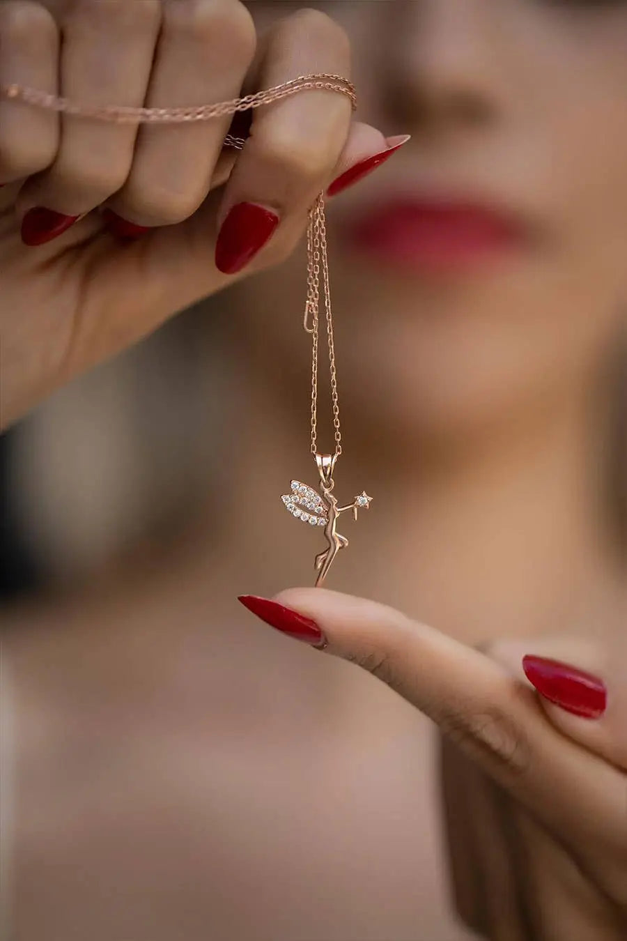 Close-up of a hand with red nail polish holding the Einzelstück - 14K Gold Plated Fairy Necklace with Black Zircona stones - Fairy Gold Necklace - Gold Necklaces Gift for Women, featuring a delicate fairy pendant with sparkling wings and a star wand.