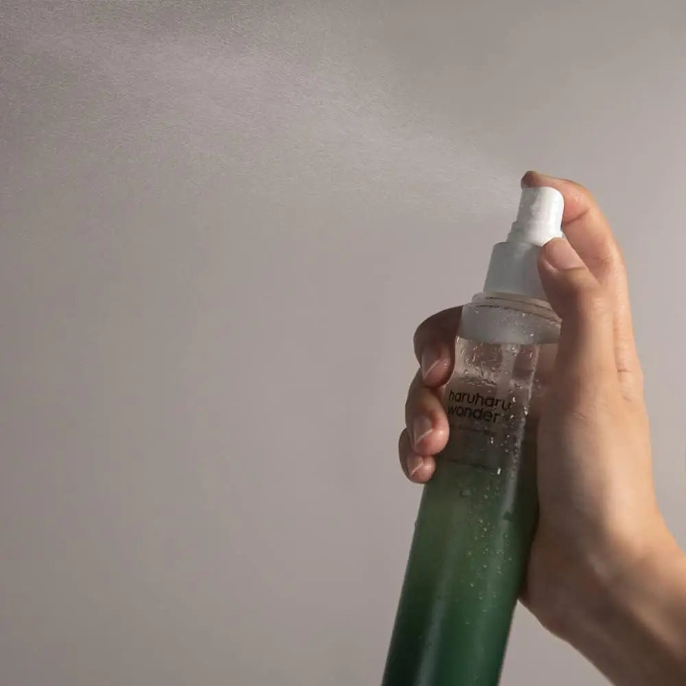 Hand holding and spraying haruharu wonder Black Bamboo Mist 150ml, releasing a fine mist of hydrating facial spray against a neutral background.