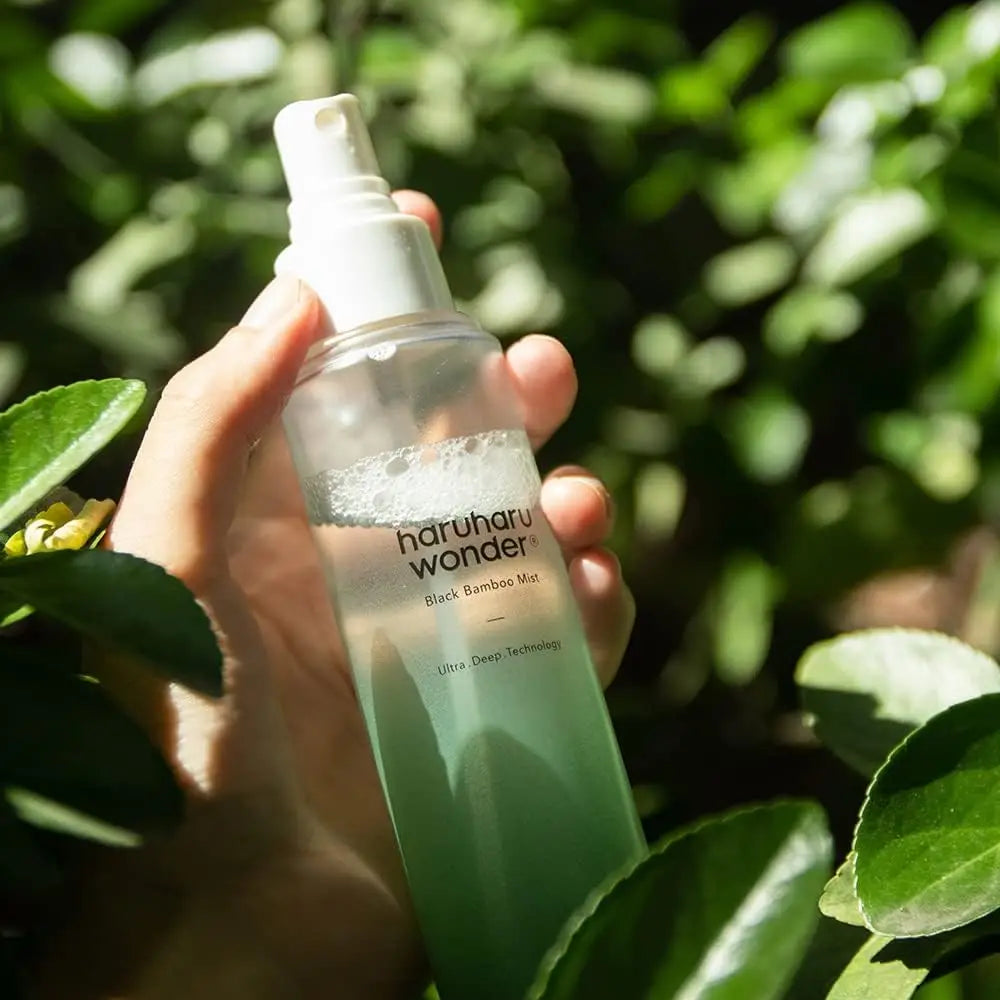 Hand holding a clear spray bottle labeled haruharu wonder Black Bamboo Mist 150ml with green liquid inside, surrounded by lush green leaves in bright natural light.