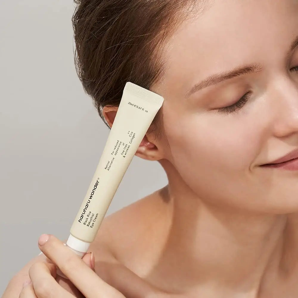 Close-up of a woman gently holding haruharu wonder Black Rice Bakuchiol Eye Cream 20ml, 2pcs tube near her face with a soft smile and closed eyes.