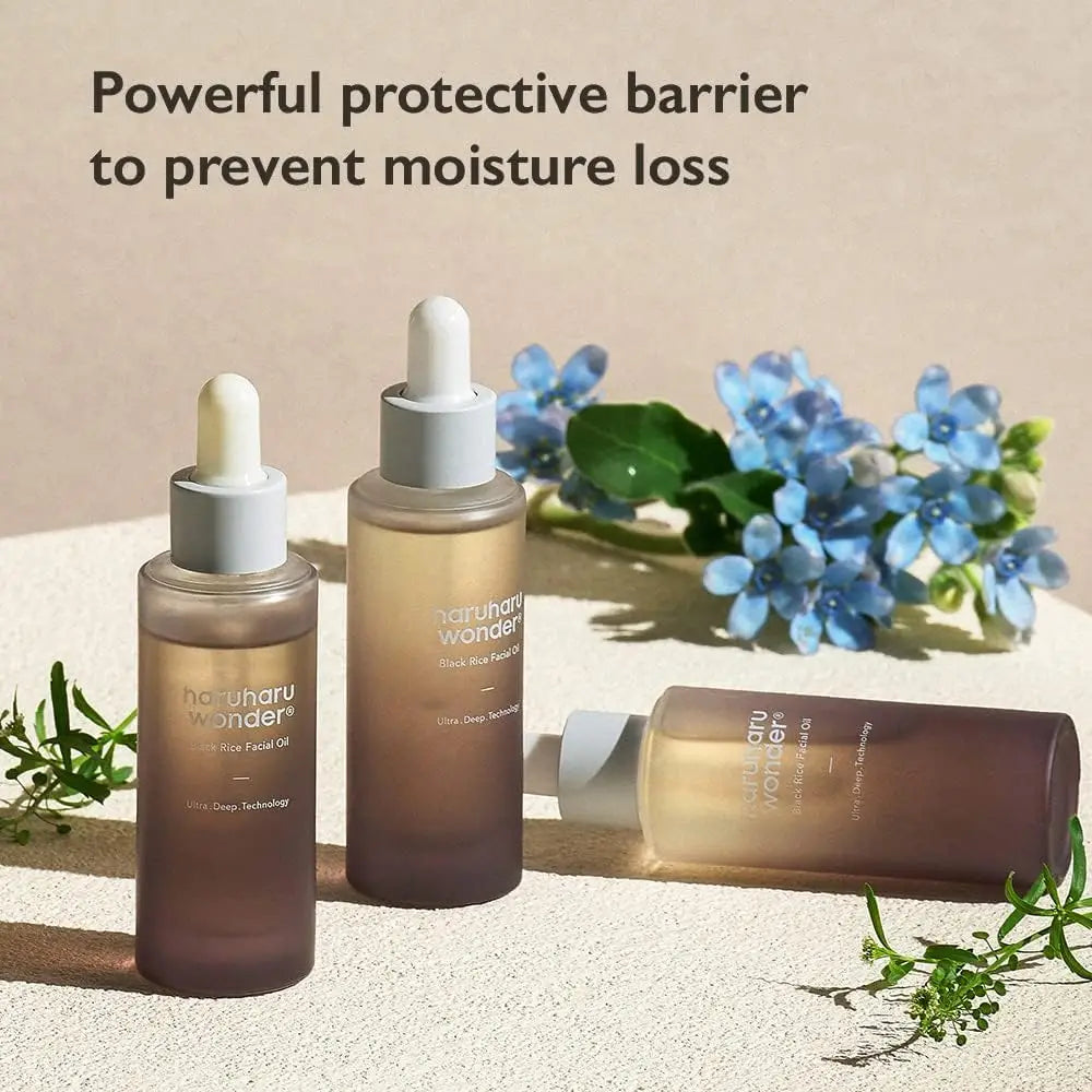 Three frosted glass bottles of haruharu wonder Black Rice Facial Oil 30ml with dropper caps arranged on a textured surface alongside green leaves and small blue flowers, emphasizing hydration and moisture protection.