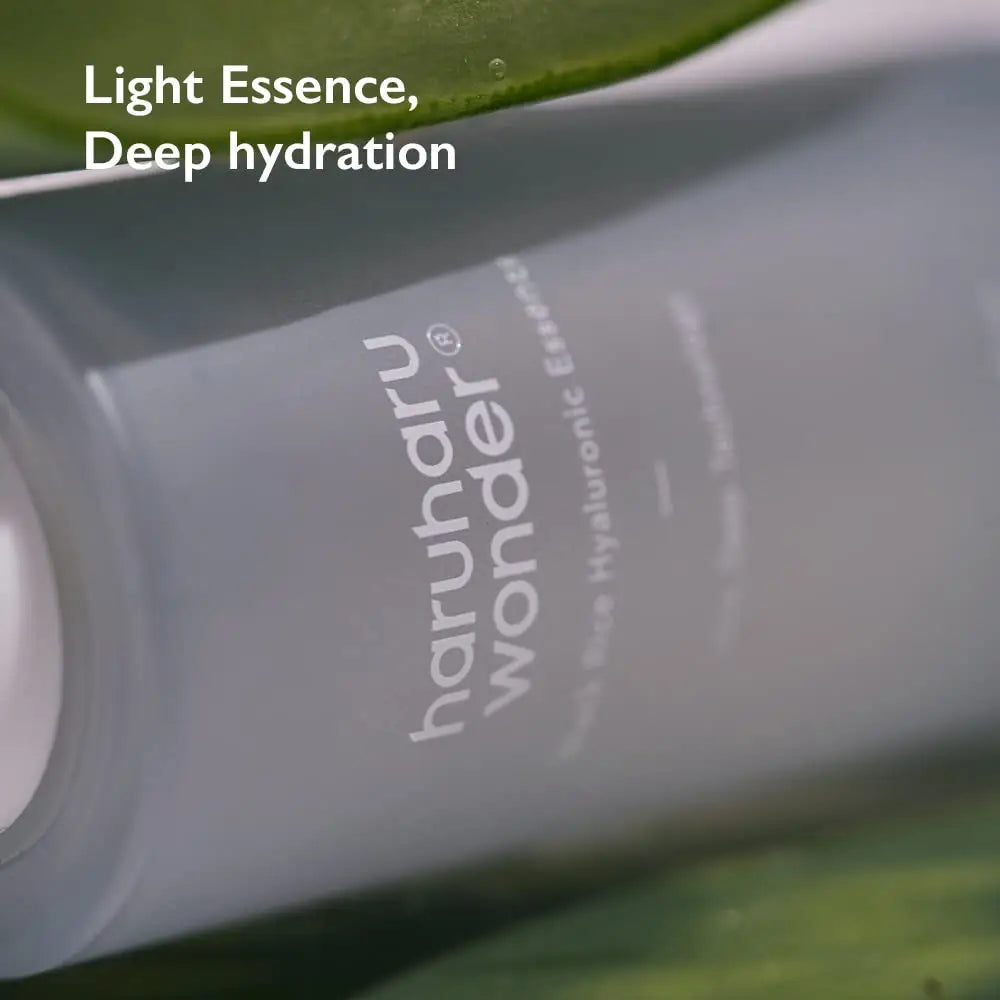 Close-up of a grey bottle of haruharu wonder Black Rice Hyaluronic Essence 50 ml resting among green leaves with text reading Light Essence, Deep hydration on the upper left.