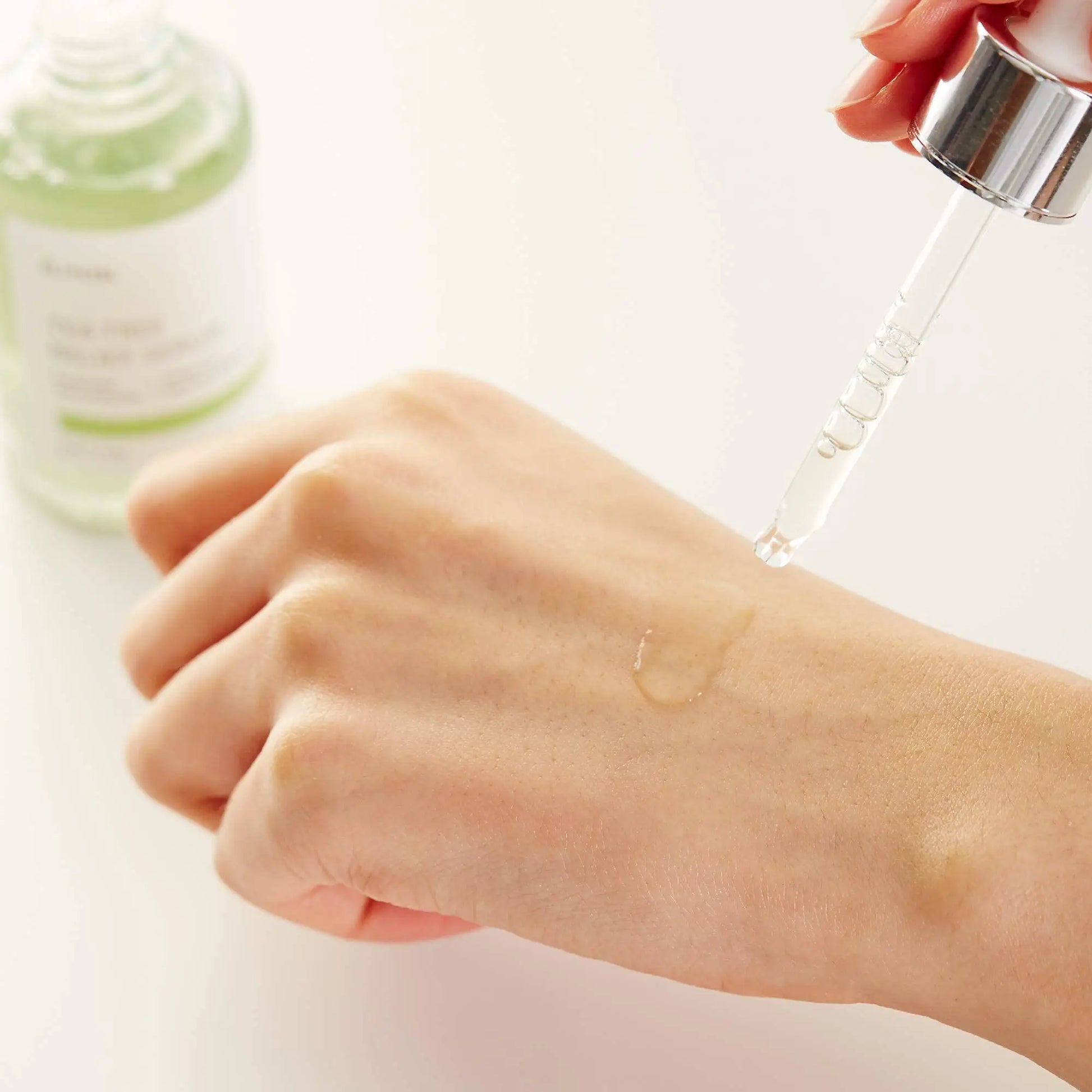 A close-up of a hand applying a drop of serum from a glass dropper with the iUNIK Beta Glucan Power Moisture Serum 50ml + iUNIK Tea tree Relief Serum 50ml bottle blurred in the background.