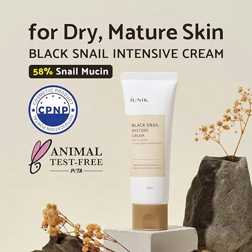 Tube of iUNIK Black Snail Restore Cream 60ml displayed on a stone pedestal with natural dried plants and rocks in the background, emphasizing its suitability for dry, mature skin and 58% snail mucin content.