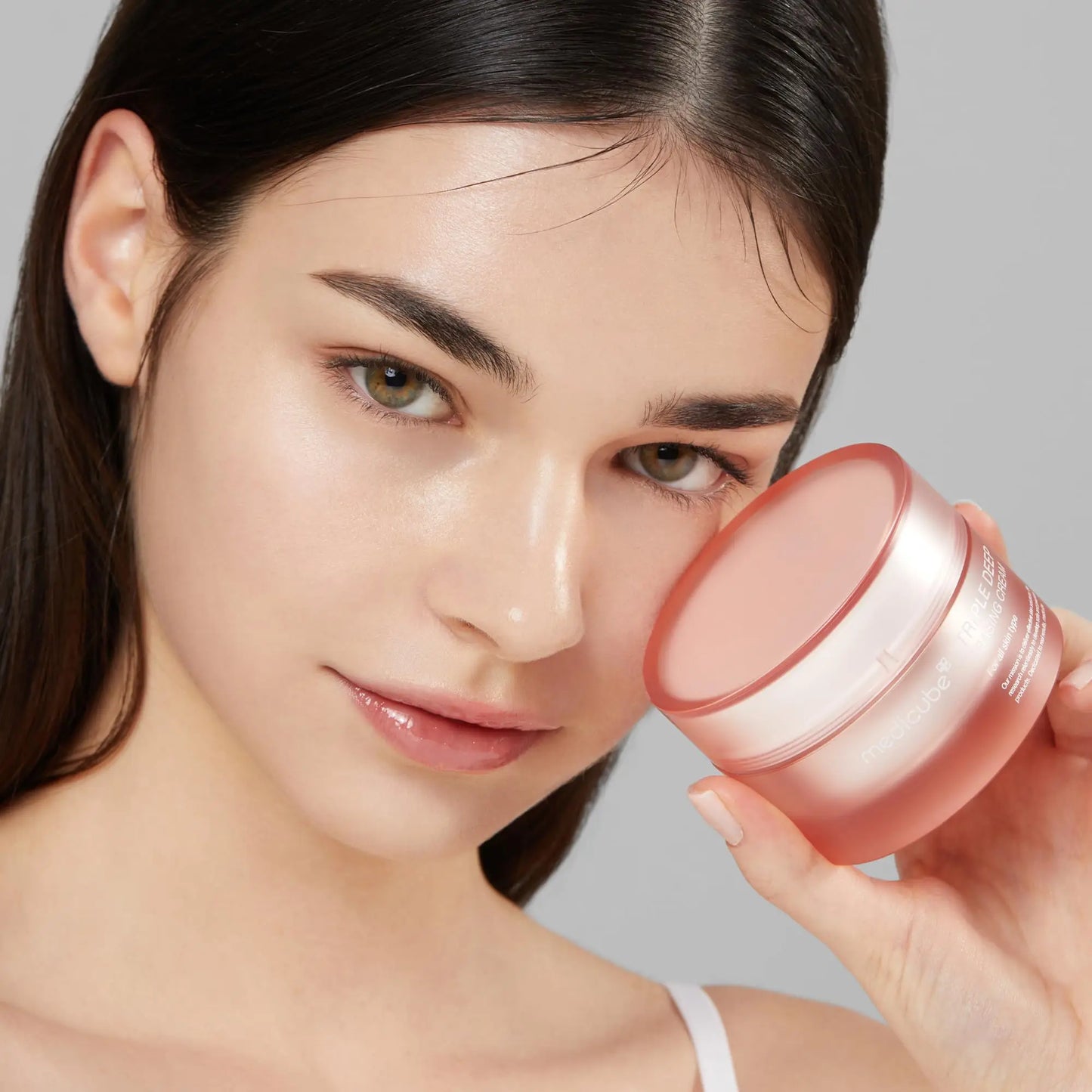 Young woman with clear glowing skin holding a jar of medicube Triple Collagen Cream 50ml near her face, showcasing hydration and radiance benefits.