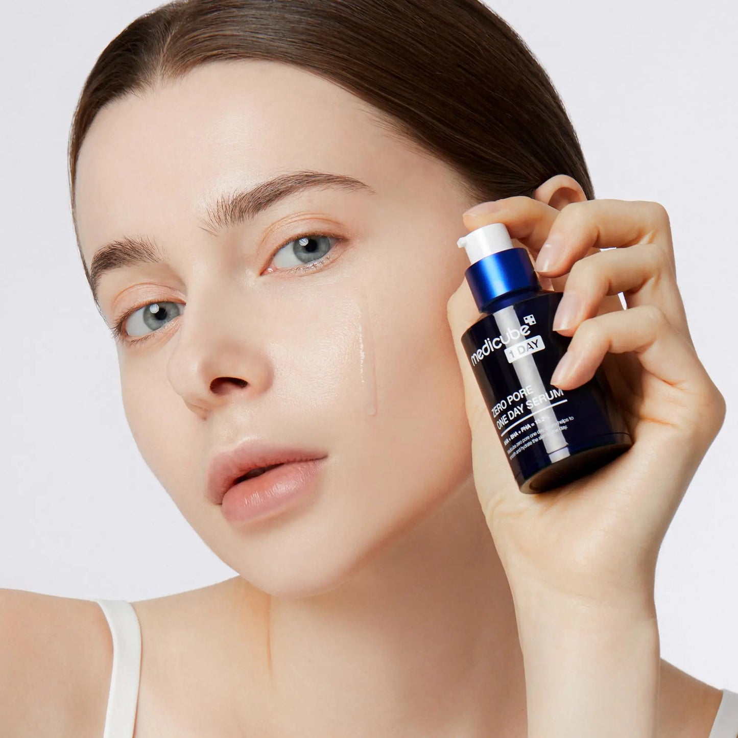 Close-up of a woman with clear skin holding a blue bottle of medicube Zero Pore Oneday Serum 30ml near her cheek with serum applied on her face.