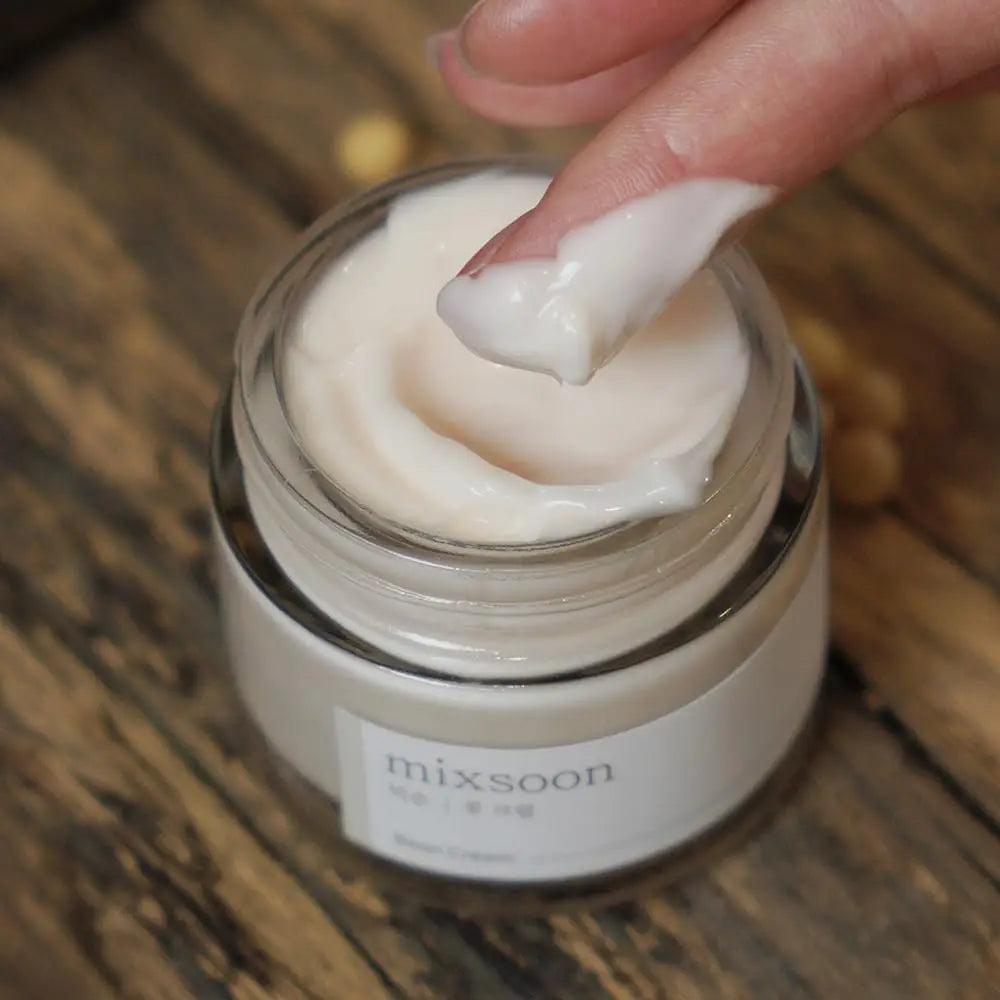 Open jar of Mixsoon Bean Cream 50ml with creamy white texture, a fingertip scooping a dollop of the facial cream on a wooden surface background.