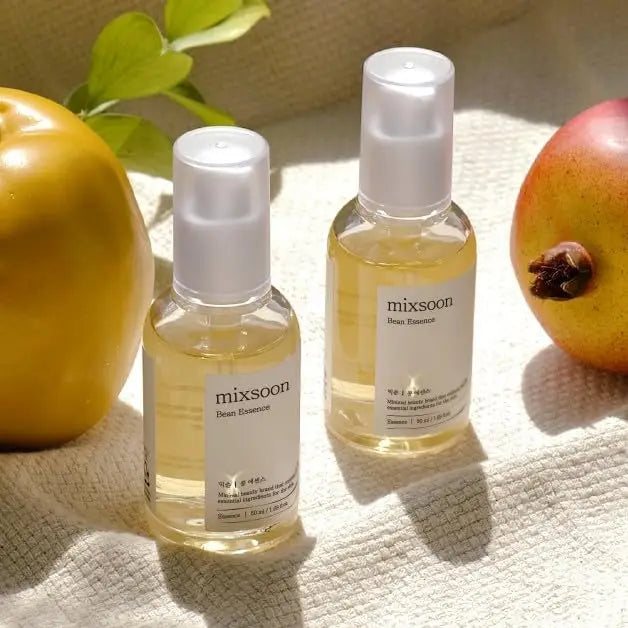 Two clear bottles of Mixsoon Bean Essence facial serum placed on a light fabric surface with natural light, surrounded by fresh fruit and greenery.