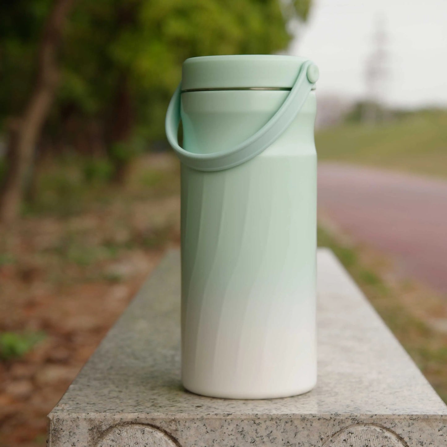 Portable Breast Milk and Water Cooler 24-Hour Extended Cooling | Ice-Free Portable Milk Storage for Travel and Work | All-in-One Ice-Free Cooler and Baby Bottle in a soft green and white design with a handle outdoors on a stone bench.