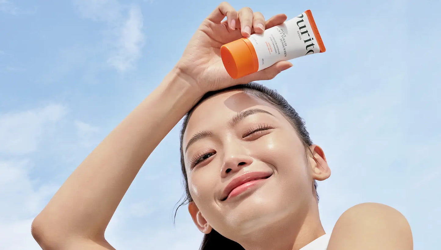 Woman smiling with glowing skin holding PURITO Daily Soft Touch Sunscreen against a clear blue sky background.