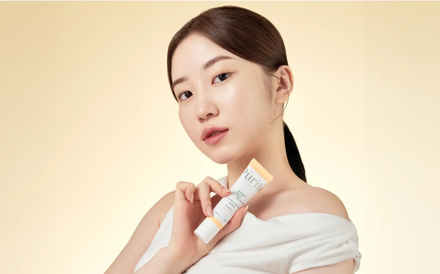 Young woman with clear skin holding a tube of PURITO Wonder Releaf Centella BB Cream 30ml against a soft beige background, showcasing natural beauty and skincare.