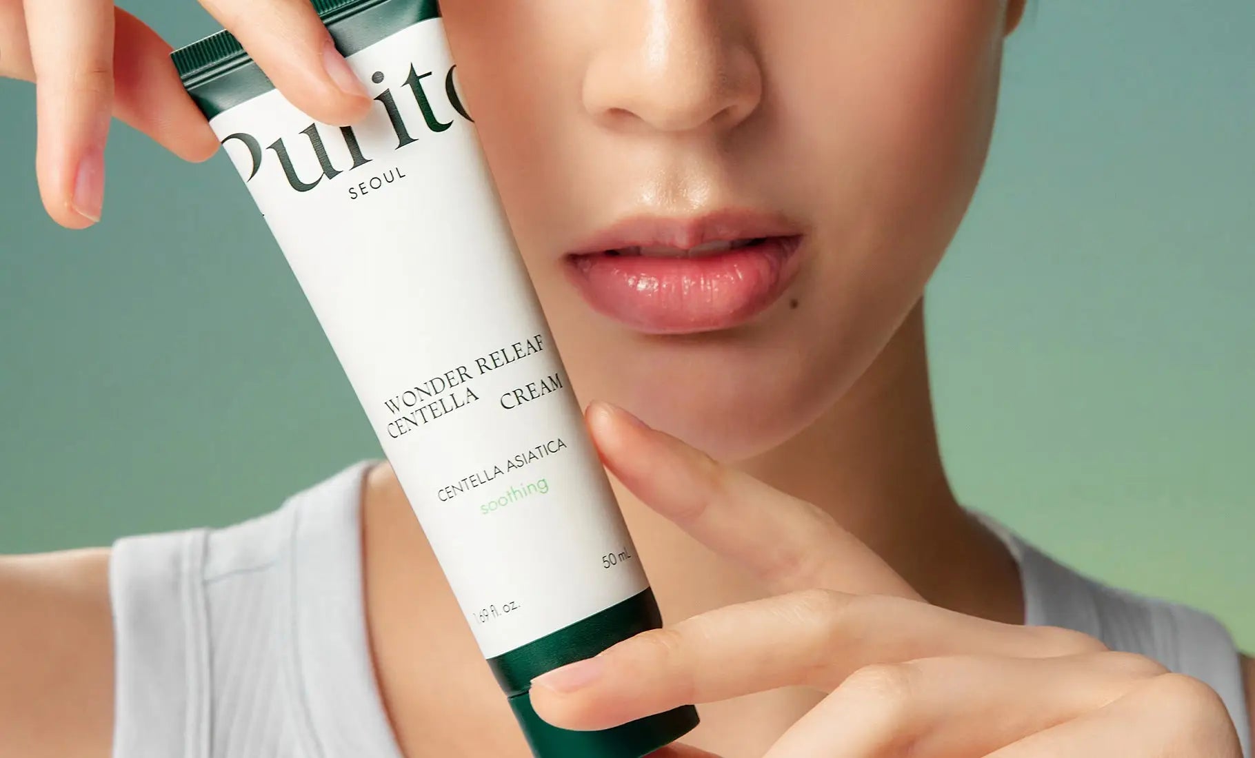 Close-up of a person holding a tube of PURITO Wonder Releaf Centella Cream 50ml near their face, showcasing smooth skin and natural makeup.
