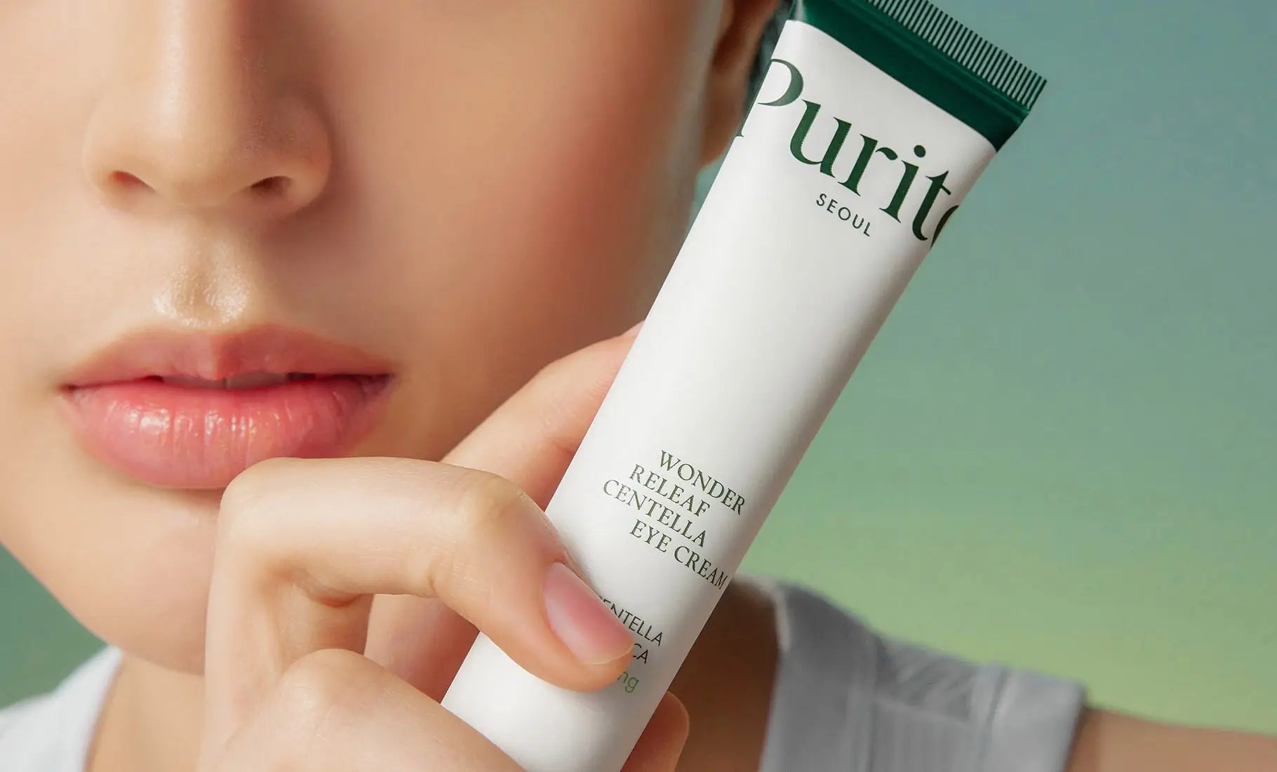 Close-up of a person holding a tube of PURITO Wonder Releaf Centella Eye Cream 30ml near their face, highlighting smooth skin and delicate application.