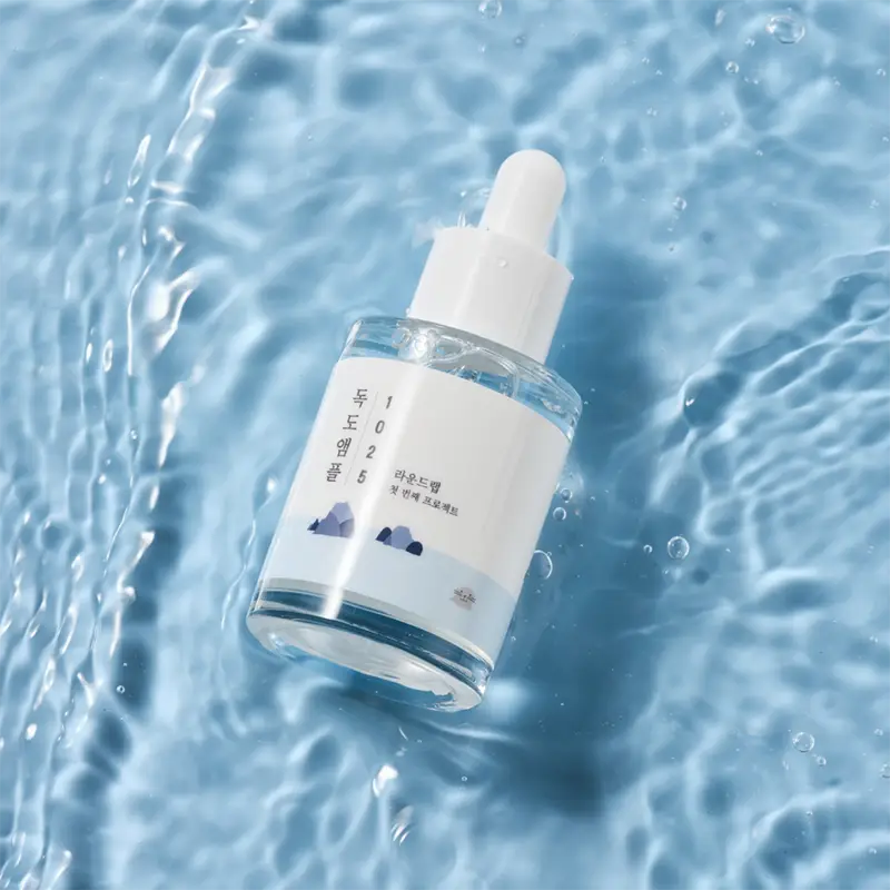 ROUND LAB 1025 Dokdo Ampoule Serum 45g in a clear glass bottle with white dropper cap, floating on rippling blue water surface, highlighting hydration and freshness.