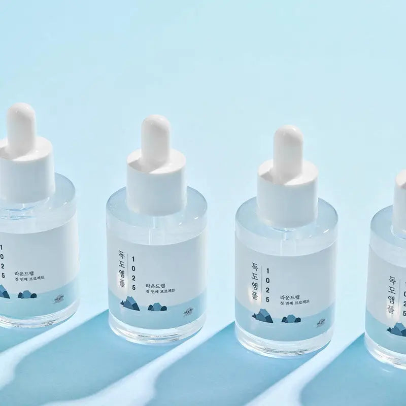 Four clear glass dropper bottles of ROUND LAB 1025 Dokdo Ampoule Serum 45g with white caps and minimalistic labels on a light blue background with soft shadows.