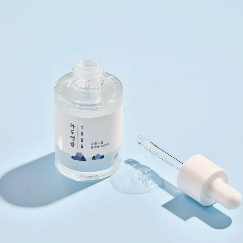 Transparent glass bottle of ROUND LAB 1025 Dokdo Ampoule Serum 45g with dropper applicator and a small amount of serum on a light blue surface under soft lighting.