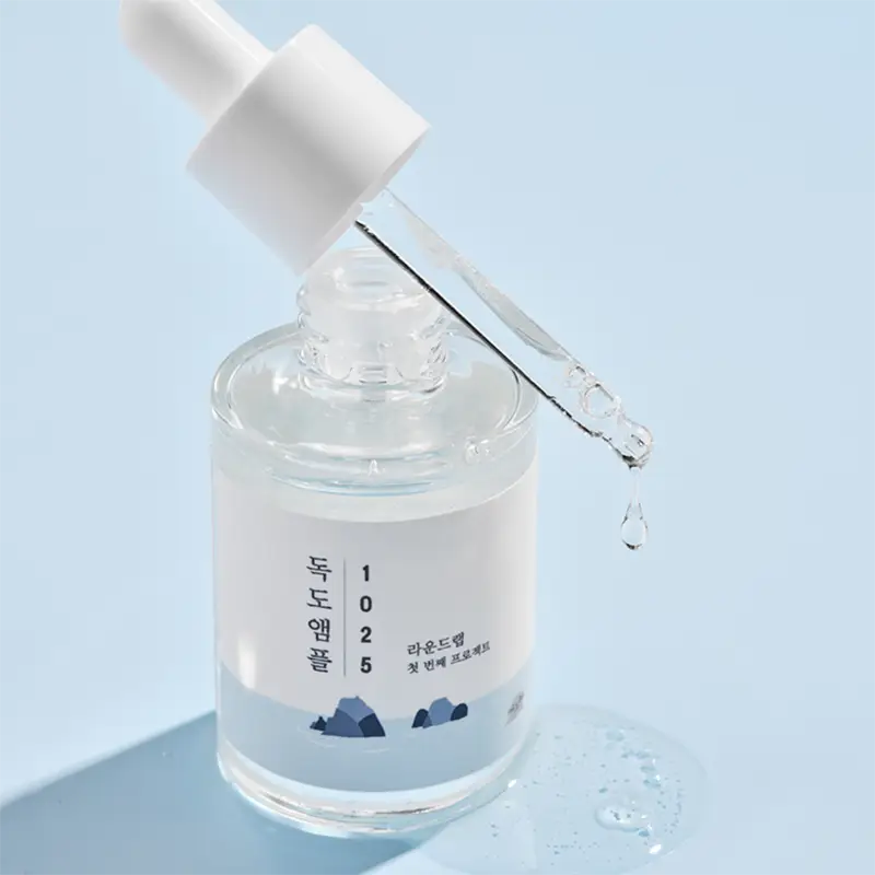 Clear serum dripping from a dropper into a glass bottle with Korean text and minimalist mountain design, showcasing ROUND LAB 1025 Dokdo Ampoule Serum 45g for skin care.