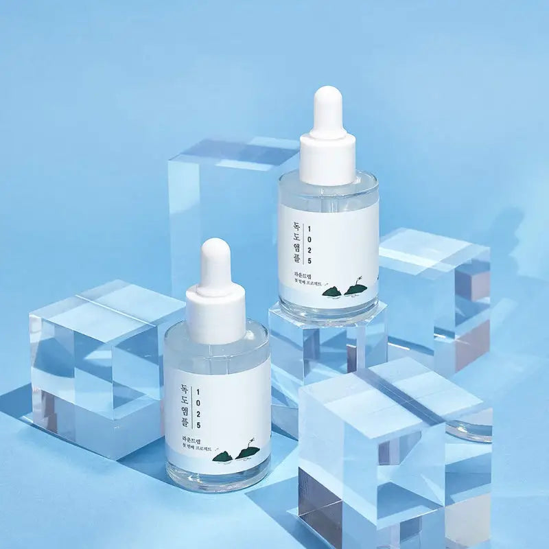 Two clear glass bottles of ROUND LAB 1025 Dokdo Ampoule Serum 45g with white dropper caps displayed on and around transparent acrylic cubes against a light blue background