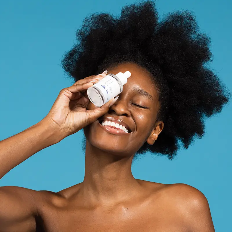 Smiling woman with natural afro hair holding a bottle of ROUND LAB 1025 Dokdo Ampoule Serum 45g near her eye against a bright blue background.