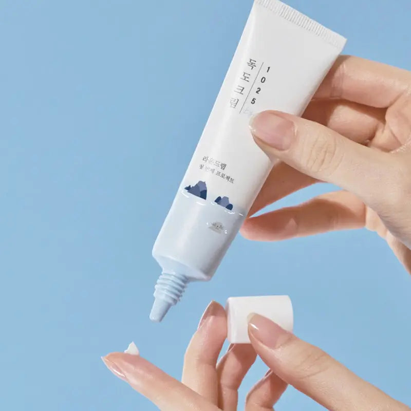 Hand holding and squeezing a white tube of Round Lab 1025 Dokdo Eye cream 30ml with a small amount of cream on fingertip against a blue background