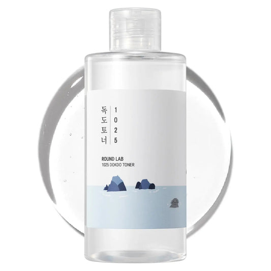 Clear plastic bottle of ROUND LAB 1025 Dokdo Toner 100ml with minimalist white label featuring ocean rocks and gentle waves illustration on a white background with translucent toner drop behind it.