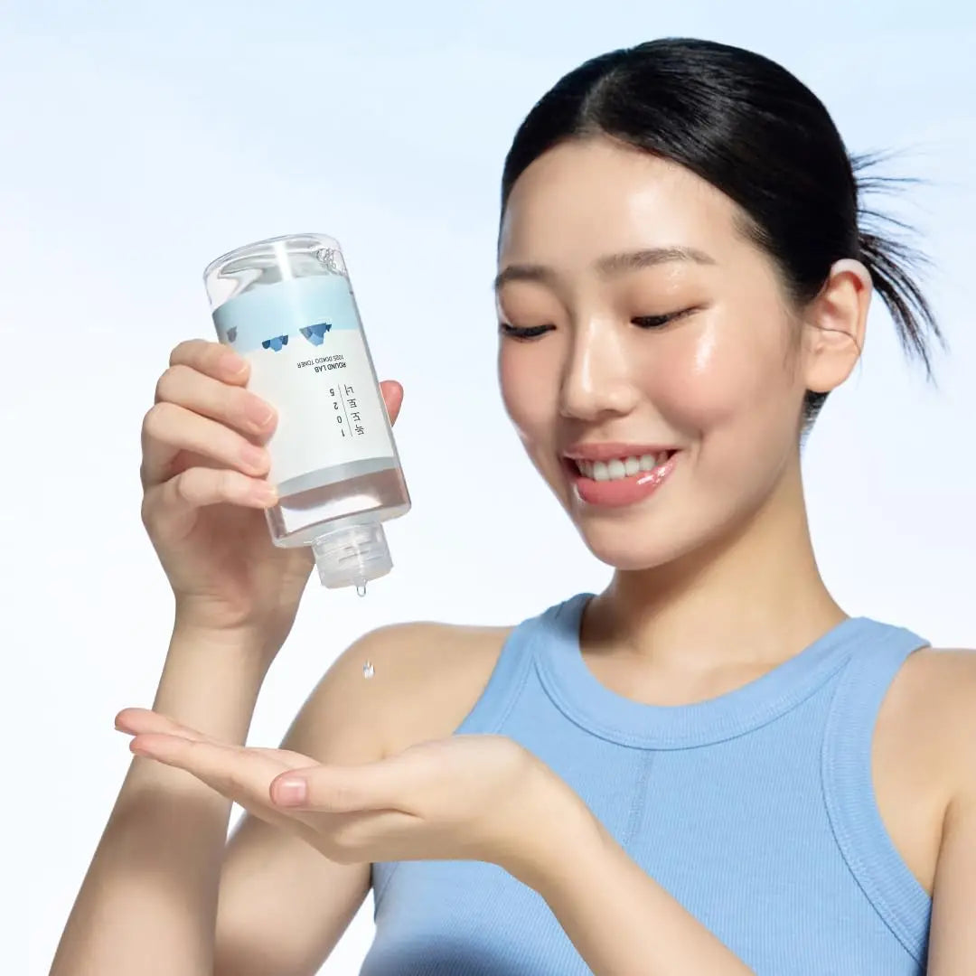 Woman wearing a blue sleeveless top smiling while pouring Round LAB 1025 Dokdo Toner 200ml + Dokdo Lotion 200ml liquid onto her hand against a clear sky background
