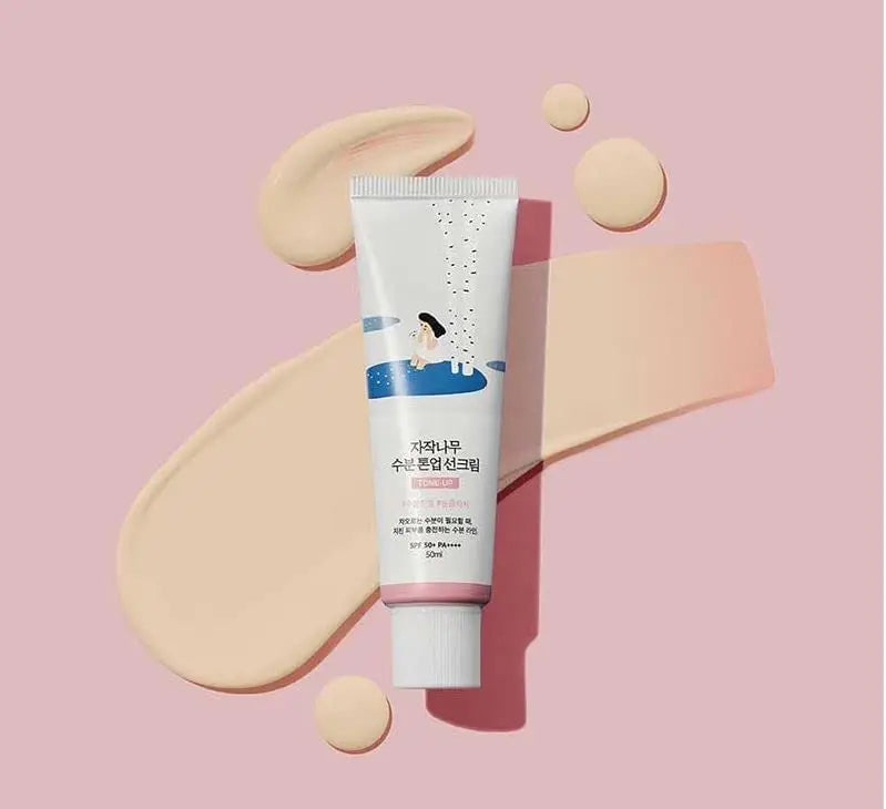 Tube of ROUND LAB Birch Juice Moisturizing Tone Up Sunscreen 50ml on a pink background with creamy beige sunscreen texture swatches showcasing smooth application and hydration.